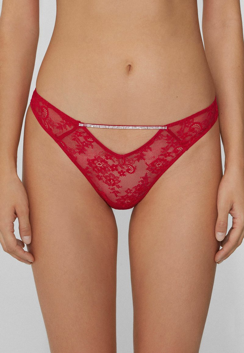 String en dentelle rouge avec un motif floral, une couverture minimale et un accent décoratif argenté le long de la ceinture. Texture douce et transparente.