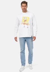 Langærmet hvid bomulds T-shirt med et farverigt SpongeBob SquarePants grafik, parret med lyseblå jeans og hvide sneakers.