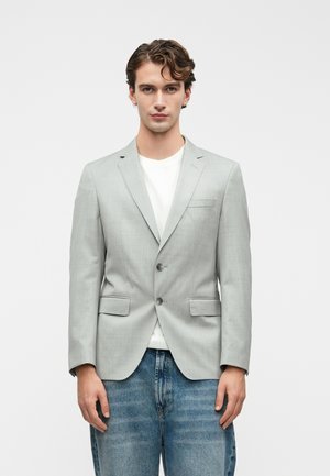 Blazer gris clair au design structuré, avec deux boutons et des revers crantés. Porté sur une chemise blanche, associé à un jean en denim bleu.