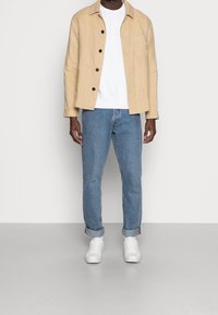 Giacca di lino beige con bottoni neri, felpa bianca, jeans blu e sneakers bianche. La giacca presenta una tasca sul petto; vestibilità casual.