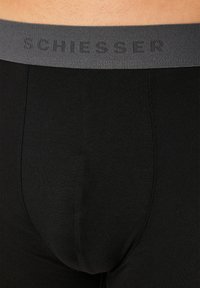 Schwarze Boxershorts mit einem grauen elastischen Bund, der mit eingeprägtem "SCHIESSER"-Text versehen ist. Glatte Textur, körperbetontes Design.