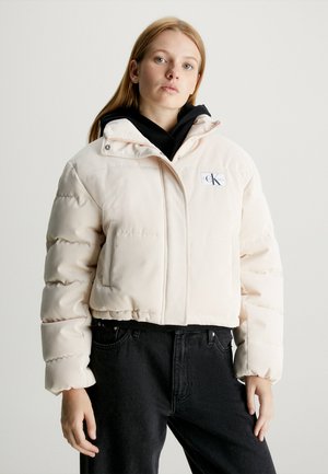 Young woman wearing a light beige puffer jacket over a black hoodie and black jeans, standing against a plain background.