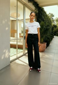 Biała koszulka z logo "TOMMY HILFIGER", ciemne, szerokie jeansy oraz bordowe sneakersy. Widok z zewnątrz z zielenią i nowoczesnym wnętrzem.