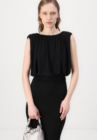 Black sleeveless top with gathered neckline and elastic waist, paired with a fitted skirt. Model holds a silver satin handbag.