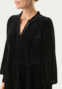 Rochie din catifea neagră cu decolteu în V, guler pliat, mâneci lungi și talie strânsă pentru o siluetă fluidă.