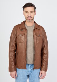 Braune Lederjacke mit Kragen, zwei Brusttaschen und Reißverschluss. Sie hat eine glatte Textur und kontrastierende Nähte. Über einem hellgrauen Pullover getragen.