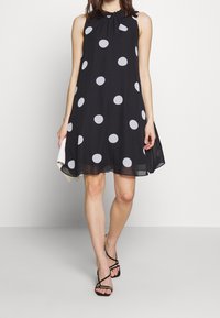 Svart ärmlös klänning med stora vita prickar, med en rynkad halsringning och en mjuk A-linjeform. Tyget är lätt och slätt.