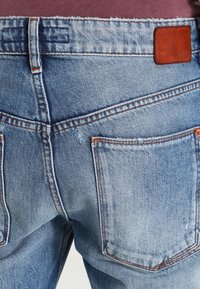 Jasnoniebieskie dżinsy z widocznymi tylnymi kieszeniami, pomarańczowym przeszyciem, szlufkami na pasek i brązową skórzaną naszywką z marką na pasku.