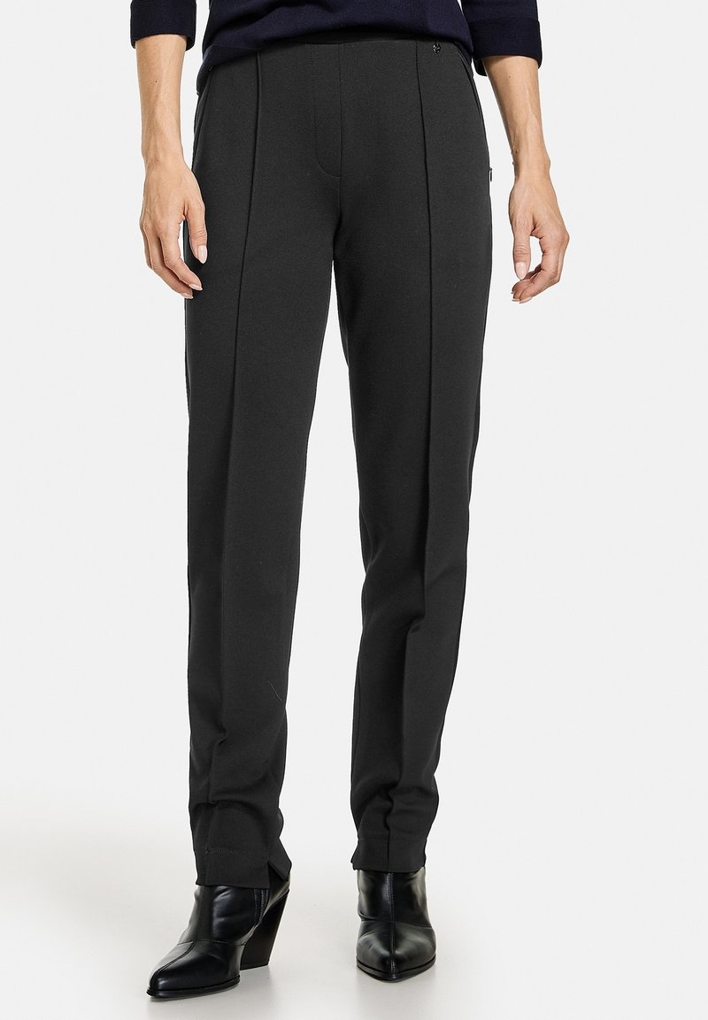Pantalons tailleur noirs à coupe droite, avec des plis avant, une texture lisse et des poches. Portés avec des bottes noires à talons.