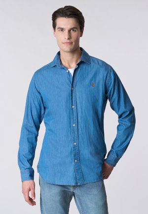 Giovane uomo con capelli scuri indossa una camicia di jeans blu abbottonata sopra una maglietta bianca e jeans azzurri, in piedi davanti a uno sfondo semplice.