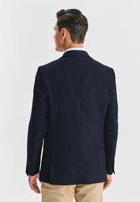 Granatowa, dopasowana marynarka z teksturowaną tkaniną, z pojedynczym rozcięciem z tyłu i guzikami na mankietach, noszona na jasnych spodniach.