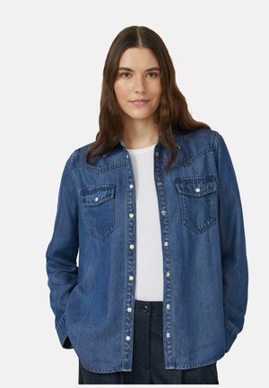 Junge Frau mit langen braunen Haaren, die ein blaues Denim-Hemd über einem weißen Oberteil und dunklen Hosen trägt, steht vor einem einfarbigen Hintergrund.
