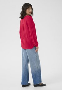Röd långärmad blus med knäppning vid manschetten, i kombination med vida blå denimjeans med en ljus till mörk ombre-effekt och svarta platta skor.