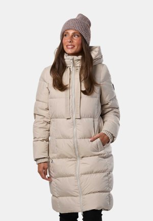Mujer que lleva un abrigo de invierno largo beige acolchado con capucha y un gorro de punto marrón claro, de pie con una mano en el bolsillo.
