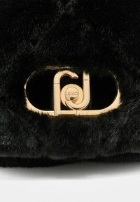 Crna torba od umjetnog krzna s zlatnim logotipom, s plišastom teksturom i pravokutnim oblikom.