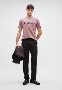 Pánske ružové tričko s nápisom "HUGO" vytlačeným tučnými čiernymi písmenami, v kombinácii s tmavými nohavicami. V jednej ruke drží tmavý sveter.