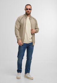 Beige gebreide zip-up vest met ribbelmanchetten en kraag, gedragen over een lichtbruin T-shirt, gecombineerd met blauwe jeans en witte sneakers.