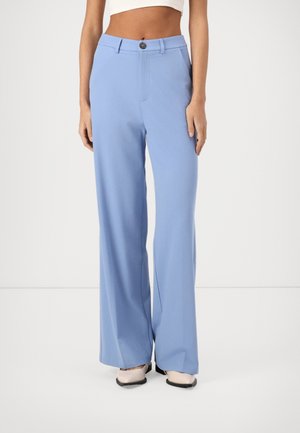 Woman wearing high-waisted, wide-leg light blue pants with a button closure, paired with beige shoes on a plain white background.