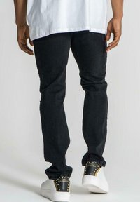 Jeans de mezclilla negra con un corte ajustado, que presentan secciones desgastadas y detalles de costura sutiles, combinados con zapatos blancos y una camisa blanca lisa.