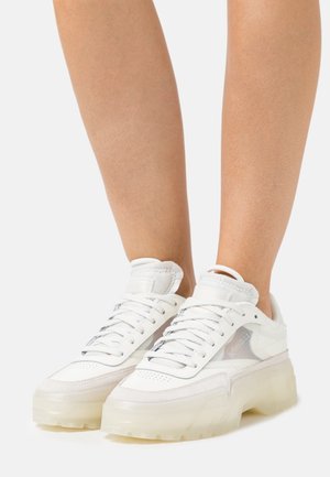 Baskets blanches avec une semelle translucide et une tige texturée. Présente des lacets, des accents en maille et un design superposé pour plus de hauteur.