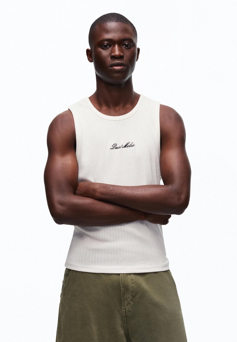 Jeune homme aux bras croisés portant un débardeur blanc avec le texte "Dust Miles" et un pantalon vert olive, debout devant un fond blanc.