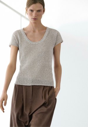 Jeune femme portant un haut tricoté gris clair à manches courtes et un pantalon marron ample, debout dans un espace intérieur lumineux et minimaliste.