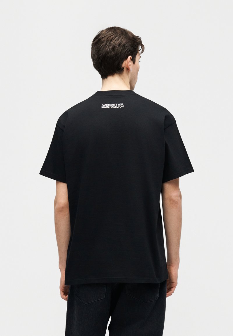 Mann trägt ein schwarzes T-Shirt mit weißem Text "Carhartt WIP Sean Hamilton" am oberen Rücken, von hinten vor einem schlichten Hintergrund gezeigt.