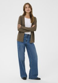 Hnědý cardigan přes bílý top, doplněný širokými modrými džíny. Oblečení má dlouhé rukávy a ploché černé boty. Jednoduchý design.