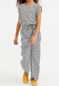 Mädchen in einem weißen Overall mit schwarzen abstrakten Punkten, Rüschenärmeln, Gürtel in der Taille und gelben Slipper-Schuhen, die vor einem weißen Hintergrund geht.