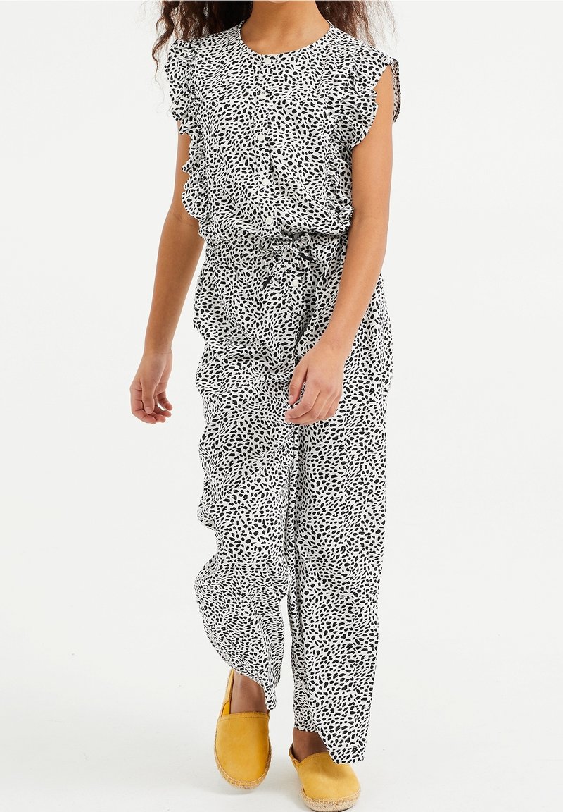Mädchen in einem weißen Overall mit schwarzen abstrakten Punkten, Rüschenärmeln, Gürtel in der Taille und gelben Slipper-Schuhen, die vor einem weißen Hintergrund geht.