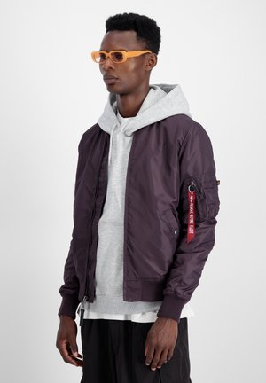 Chaqueta bomber de nailon morada con puños acanalados, combinada con un sudadera con capucha gris y gafas rectangulares naranjas. Presenta un detalle de etiqueta roja.
