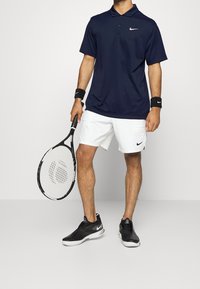 Polo Nike bleu marine, shorts blancs, chaussures Nike noires, tenant une raquette de tennis noire avec un tamis à motifs. Comprend des bracelets noirs.