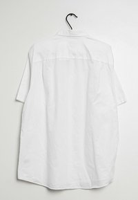 Camisa blanca de manga corta con un cuello clásico, fabricada en algodón suave y con un dobladillo redondeado, exhibida colgando de un gancho negro.