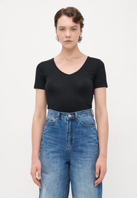 Junge Person mit kurzen, welligen Haaren, die silberne Creolen trägt, ein enges schwarzes V-Ausschnitt-T-Shirt und hoch taillierte blaue Jeans vor weißem Hintergrund.