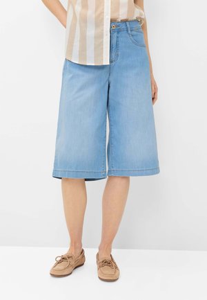 Person iført knælange lyseblå denimshorts, beige og hvide stribede skjorte og lysebrune snøreløse mokkasinsko.