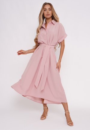 Kobieta w jasnoróżowej, midi sukience z paskiem, z krótkimi rękawami, w beżowych sandałach na wysokim obcasie, pozująca na jednolitym tle.