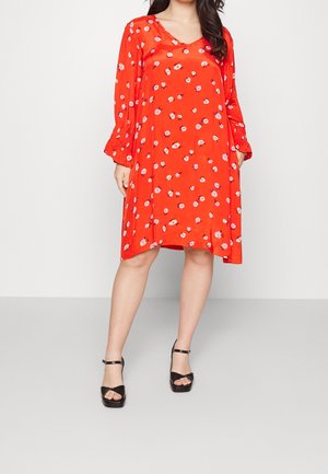 Woman wearing a loose-fitting red dress with small white and purple floral prints, long sleeves with ruffled cuffs, and black open-toe heels.