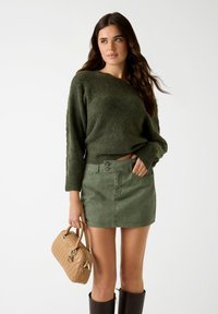 Kobieta ubrana w ciemnozielony sweter dzianinowy i pasującą mini spódniczkę, trzyma beżową pikowaną torebkę i nosi ciemnobrązowe botki do kolan.