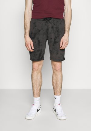 Mężczyzna ubrany w czarne szorty ze sznurkiem, białe skarpetki Nike oraz białe buty sportowe Nike, stojący na jednolitym tle.