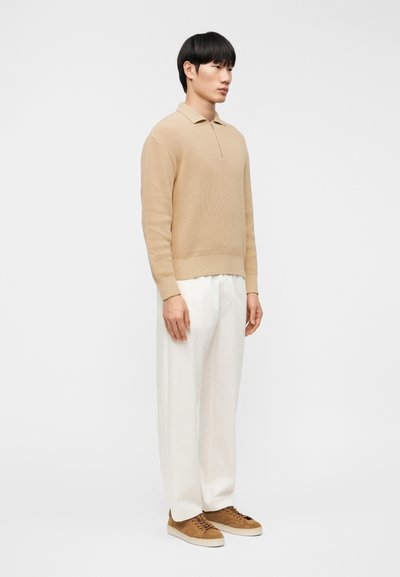 Άνδρας μοντέλο σε όρθια πλευρική στάση, φορώντας μπεζ πλεκτό που λόβερ με μακριά μανίκια, λευκό φαρδύ παντελόνι και καφέ σουέντ αθλητικά παπούτσια.
