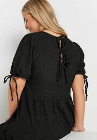 Svart klänning med texturerad blommönster, korta puffärmar, snörning i ryggen och en tierad nederkant.