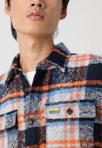 Camicia di flanella con motivo a quadri blu, arancione e grigio; presenta due tasche sul petto, chiusure a bottoni e un piccolo badge decorativo.