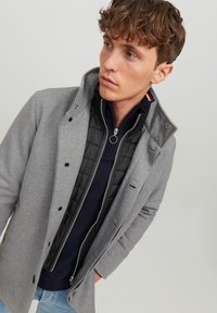 Grå uldblandet frakke med krave, der har et sort quiltet indre lag. Designet inkluderer trykknapper og en lynlåslukning foran.