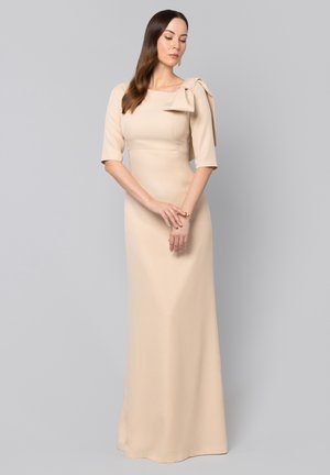 Kobieta w beżowej, sięgającej do ziemi sukni z rękawami do łokcia i dużą kokardą na ramieniu, stojąca z rękami splecionymi na tle w kolorze szarym.