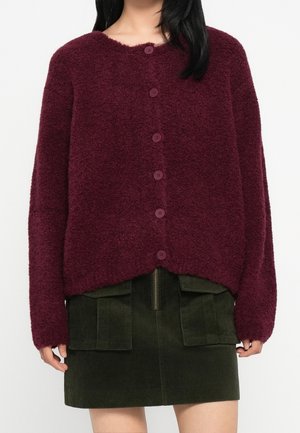 Frau trägt eine burgunderrote, geknöpfte, flauschige Strickjacke, kombiniert mit einem grünen Cord-Minirock mit aufgesetzten Vorderklappentaschen und einem zentralen Reißverschluss.