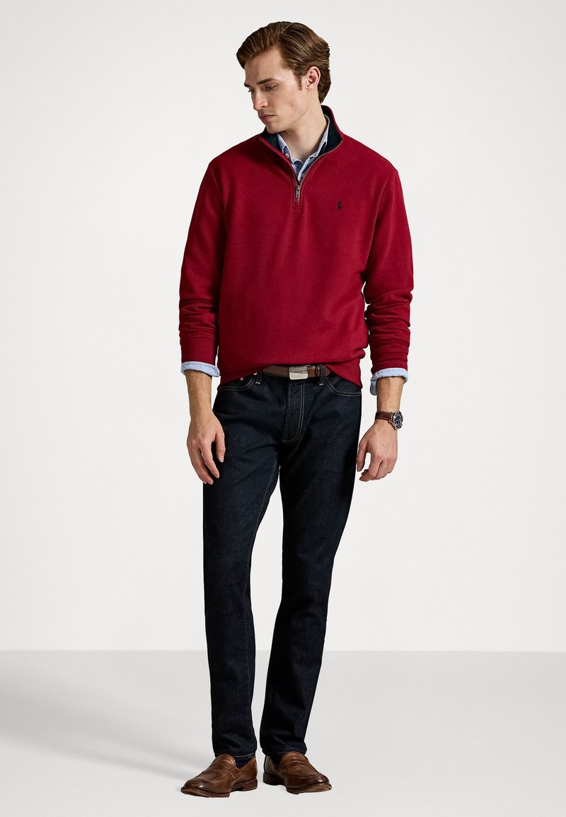 Uomo che indossa un maglione rosso con zip quarti sopra una camicia blu, jeans scuri, cintura e scarpe marroni, in piedi contro uno sfondo bianco uniforme.