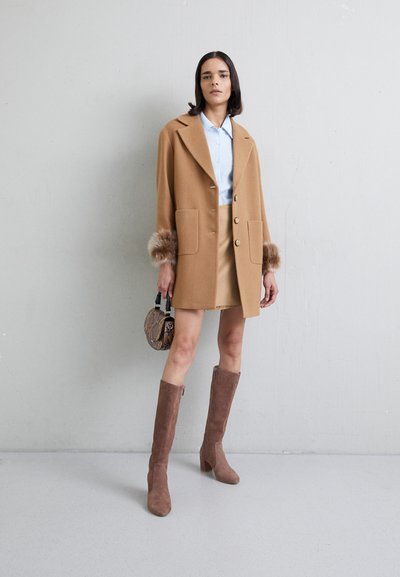 Cappotto color cammello con maniche in pelliccia sintetica, chiusura a bottoni, camicia azzurra, gonna beige, stivali marroni fino al ginocchio e una piccola borsa a tracolla con motivo.