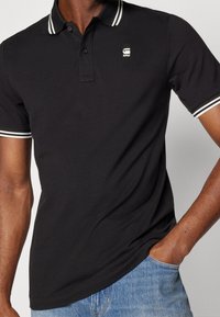 Černé polo triko z bavlny s bílými proužky na límci a okrajích rukávů. Malé logo na hrudi. Nošeno s modrými džíny.