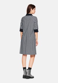 Schwarz-weiß gemustertes Kleid mit Kragen und kurzen Ärmeln, gerade geschnitten und mit seitlichen Taschen. Kombiniert mit grauen Ankle Boots.