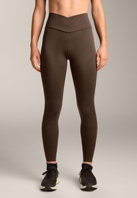 Barna, magas derekú leggings V-alakú derékkal, sima felülettel és testhezálló kialakítással, fekete sportcipőkkel párosítva.